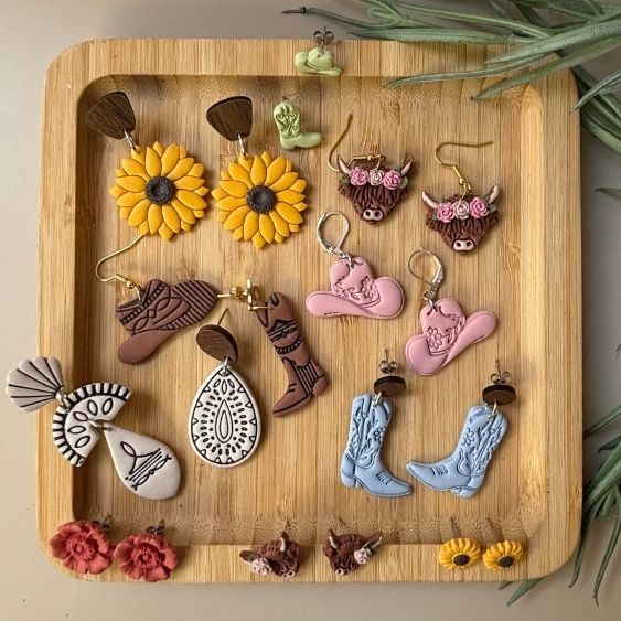 A collection of handmade western-themed dangle earrings displayed on a rectangular wooden tray. The earrings feature sunflower, cowboy boot, horseshoe, teardrop, and cowgirl hat designs in yellow, brown, pink, blue, white, and red.