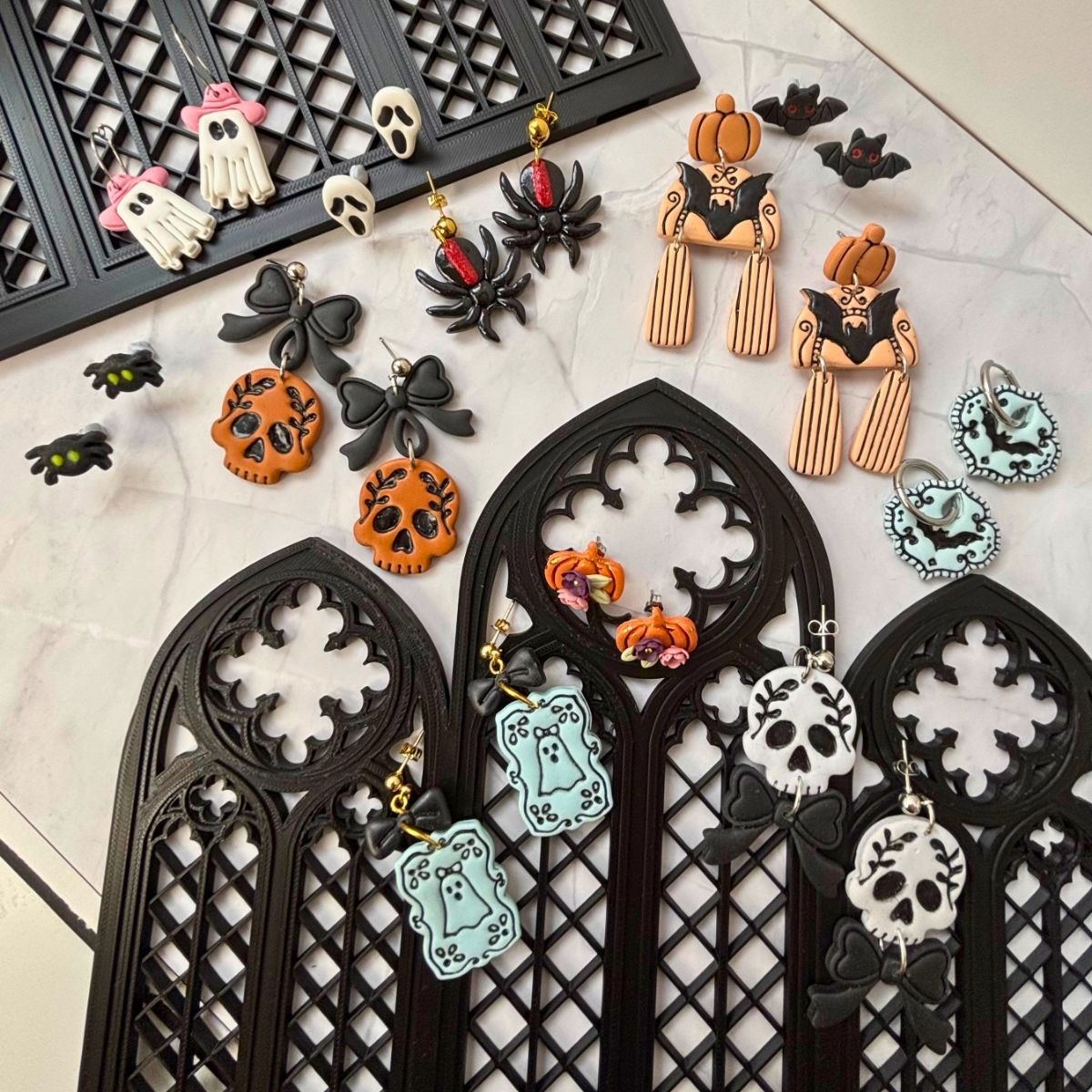 Collection of Halloween-themed earrings on black decorative arch stands. Earrings include skull, ghost, spider, bat, pumpkin, and bow motifs in black, orange, white, and pale blue,