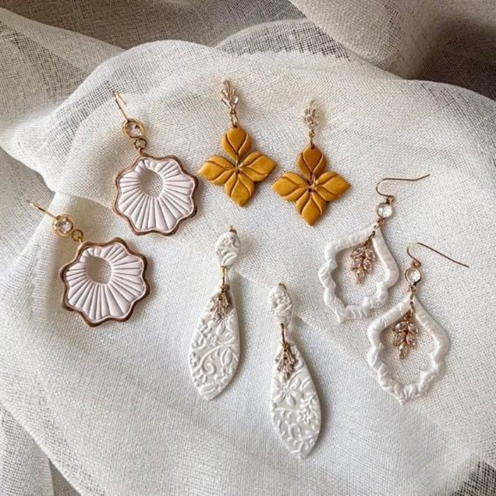 Six bohemian-style handmade earrings arranged on a textured white fabric background. The set includes gold clay flower earrings, white textured shell earrings, white teardrop earrings with gold filigree, with CZ accents. Special occasion bridal styles