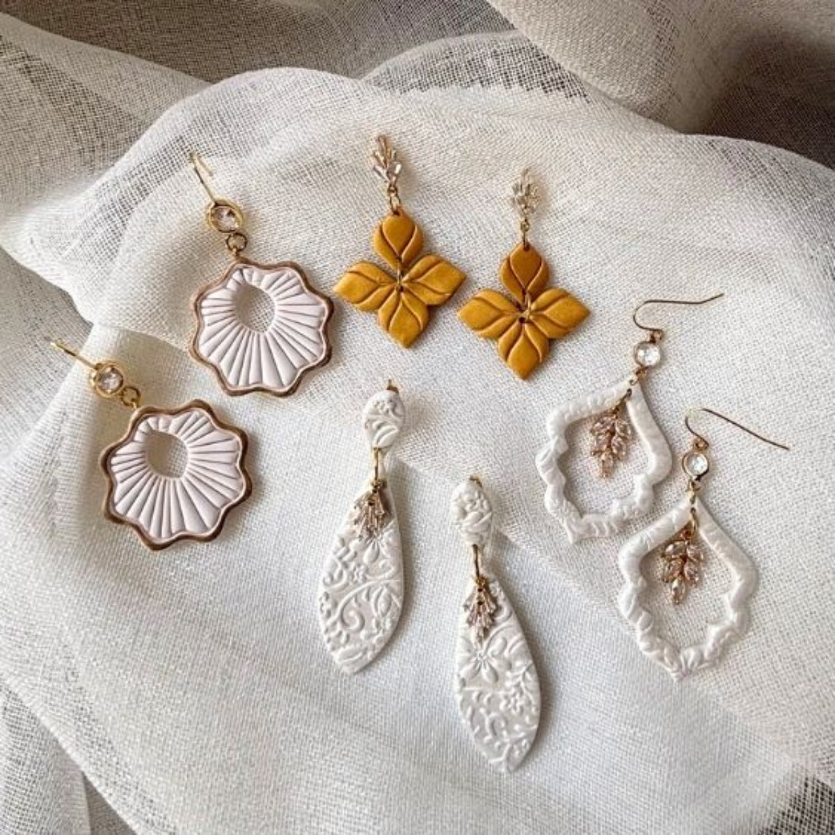 Six bohemian-style handmade earrings arranged on a textured white fabric background. The set includes gold clay flower earrings, white textured shell earrings, white teardrop earrings with gold filigree, with CZ accents. Special occasion bridal styles