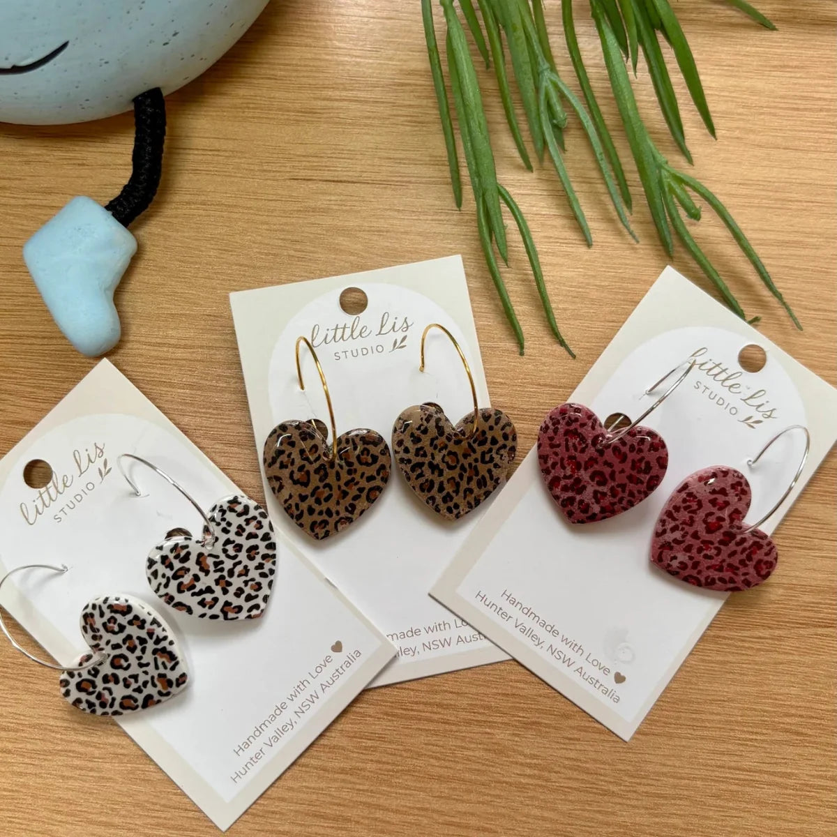Three pairs of heart-shaped leopard print earrings on display cards on a wooden tabletop. The earrings feature leopard patterns in brown, black, pink and beige, 