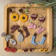 A collection of handmade western-themed dangle earrings displayed on a rectangular wooden tray. The earrings feature sunflower, cowboy boot, horseshoe, teardrop, and cowgirl hat designs in yellow, brown, pink, blue, white, and red.