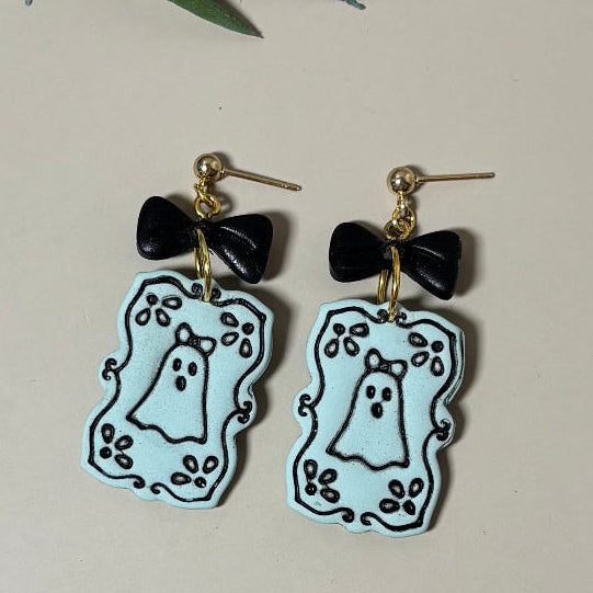 A pair of dangling earrings with black bow-shaped charms and light green ghost-shaped drops outlined in black, perfect for spooky Halloween fun