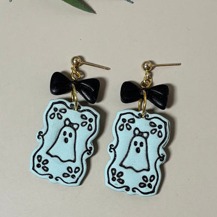 A pair of dangling earrings with black bow-shaped charms and light green ghost-shaped drops outlined in black, perfect for spooky Halloween fun