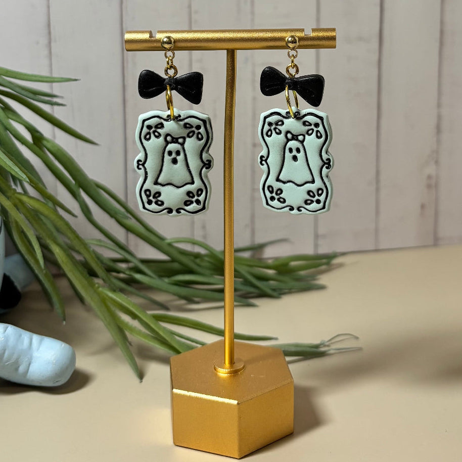 A pair of dangling earrings displayed on a gold stand. Each earring has a small black bow above a green rectangular frame with a black decorative border and a ghost illustration in the center.