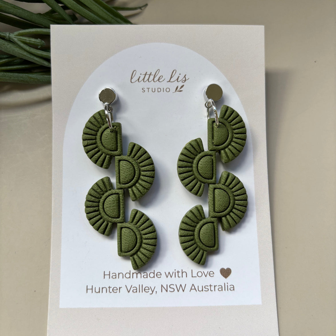 A pair of olive green textured dangle earrings displayed on a white card by Little Lis Studio photographed against a light beige background with a small green plant leaf in the corner.