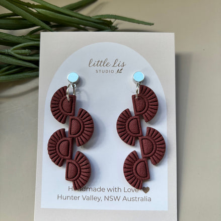 A pair of dangling earrings displayed on a white card. The earrings have a dark reddish-brown or rosewood textured geometric design with silver studs.