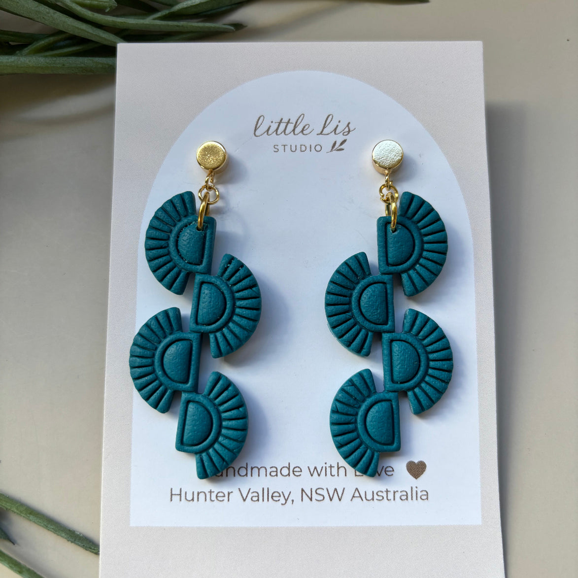 A pair of teal and gold geometric dangle earrings displayed on a white backing card that reads 'little Lis STUDIO NZ' and 'Handmade with Love, Hunter Valley, NSW Australia'. The card is styled on a neutral beige surface with green leaves in the background.