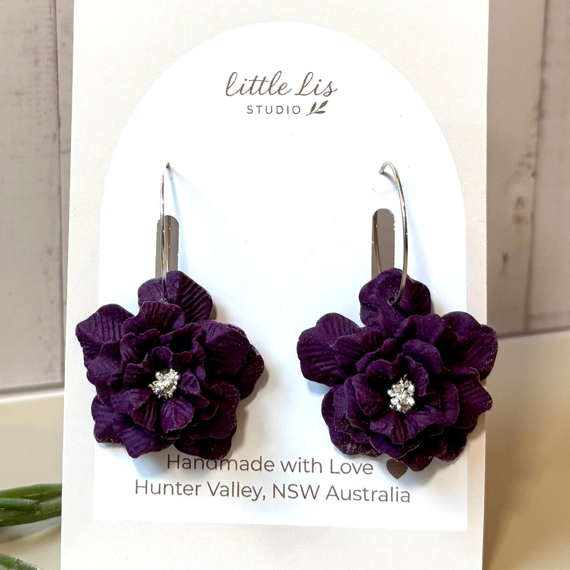 A pair of handmade dark purple polymer clay flower earrings with fine silver hoops and a small silver center, displayed on a white 'little Lis STUDIO' card.