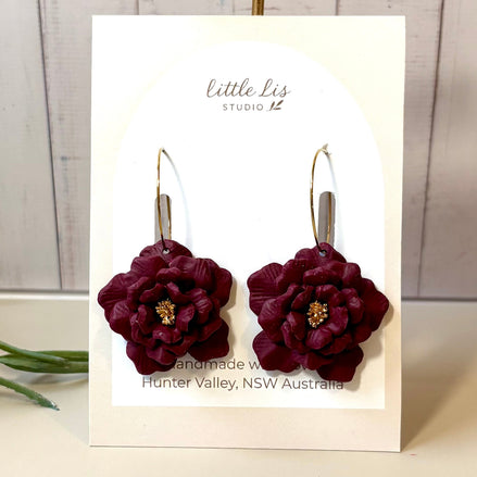 A pair of handmade dark burgundy flower drop earrings with gold hooks, displayed on a white card labeled 'little LIs STUDIO 12' against a light wood paneled background.