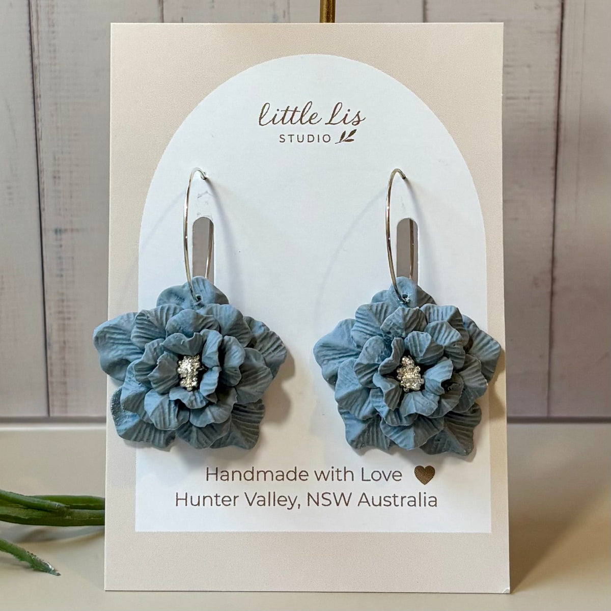 A pair of handmade dangle earrings by Little Lis Studio, featuring textured dusty blue polymer clay flowers with a small silver center and thin silver-tone hoops, displayed on a white card.