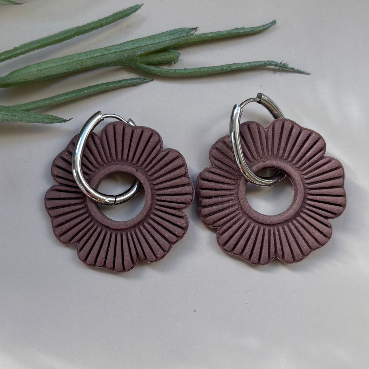 Pair of Mocha Brown flower shaped earrings with silver hoops on a light background with green leaves.